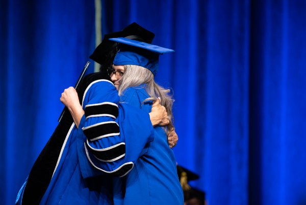 A person in a cap and gown hugs a person wearing academic regalia on stage. 