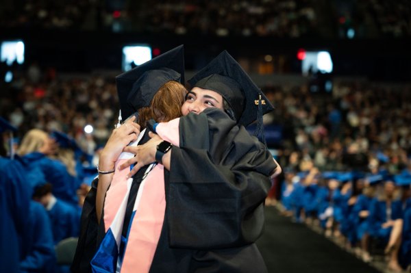A person hugs another person, both wearing caps and gowns, with a crowd behind them in an arena.