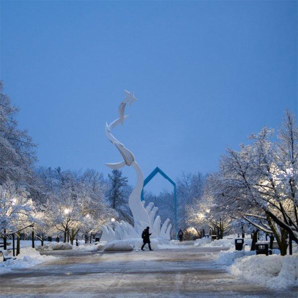 A person walks past a sculpture on a snowy college campus at dawn.
