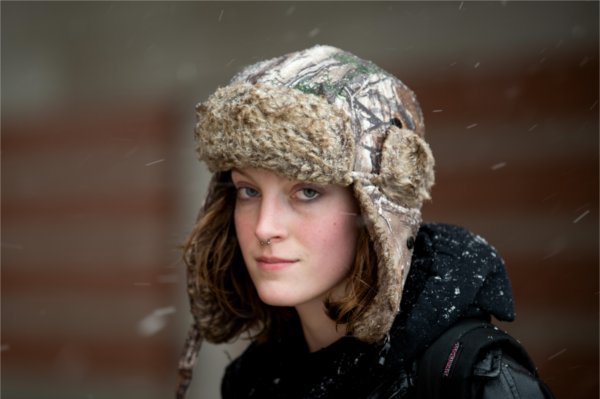 A college student looks at the camera while wearing a warm winter hat in the snow.  