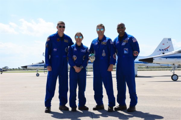 Four astronauts wearing sunglasses stand before a NASA vehicle.