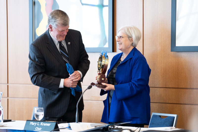 Thomas J. Haas accepts a clock from board chair Mary Kramer. The clock looks like a ship's wheel and is gold. Haas smiles brightly as he accepts the award.