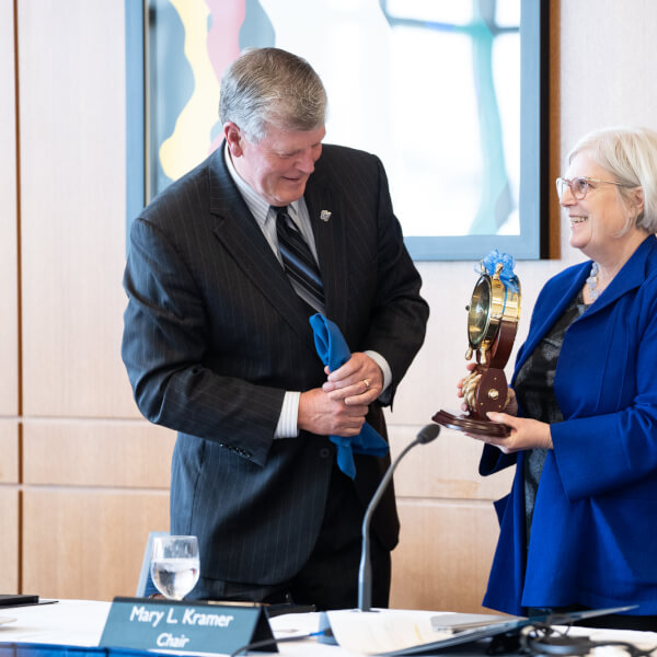 Thomas J. Haas accepts a clock from board chair Mary Kramer. The clock looks like a ship's wheel and is gold. Haas smiles brightly as he accepts the award.