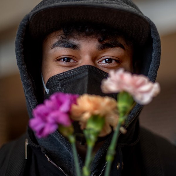 A person, wearing a mask and holding carnations, gazes into the camera.