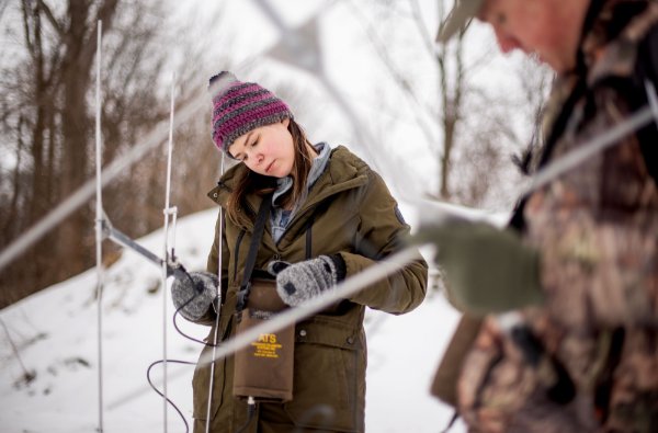 Two students hold metal devices in their hands while they stand in the snowy woods. One student is looking down at a device she has hanging around her neck. 