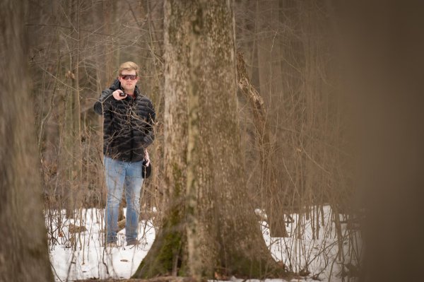 A person stands in the forest between trees pointing a devise with a red laser at a tree. The person is wearing a black coat and dark protective glasses.