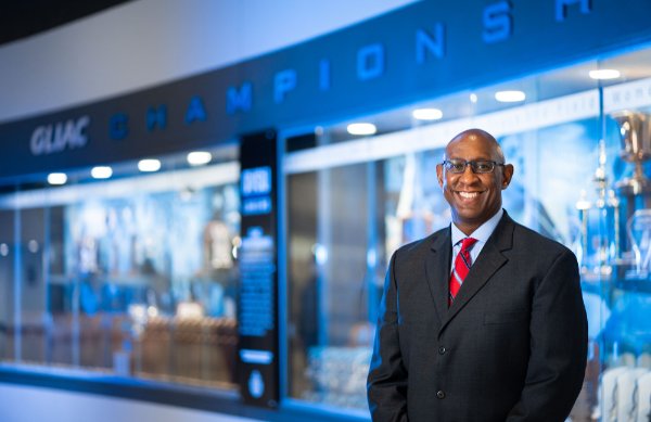 A man stands in front of a blue trophy case with the words "GLIAC Champions" above the case. 