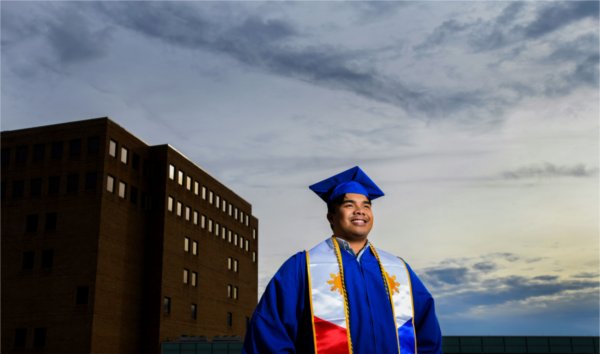 A person in a graduation cap and gown looks off into the distance with a building behind them.