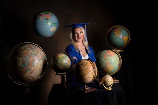 A person in a graduation cap and gown stands in a posed portrait with globes surrounding them.