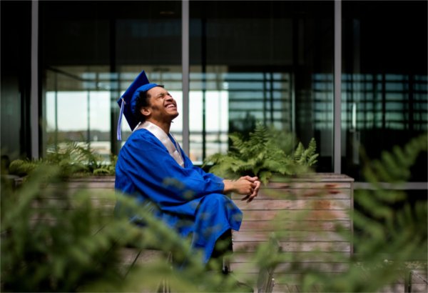 A person seated and wearing a graduation cap and gown looks upward and smiles with eyes closed amid containers filled with plants.