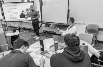 A man stands at the front of a classroom pointing to a large screen displaying a slide titled &#8220;Problem Questions,&#8221; while several students seated around a table take notes and engage with the lesson.