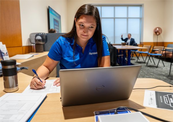A woman wearing a blue Fastenal polo shirt sits at a desk, focused on writing on a form while working on a laptop.