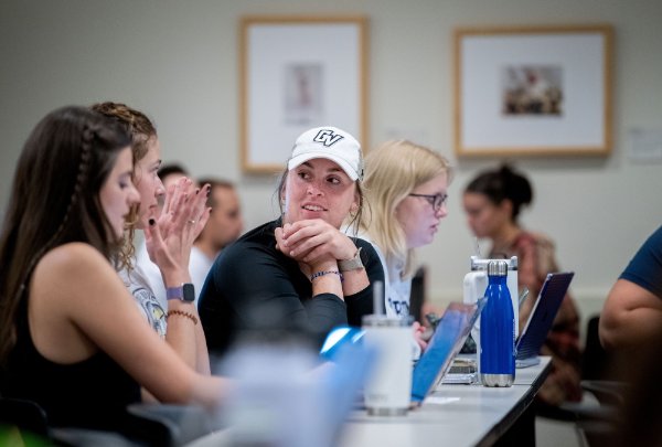 Students gesture with their hands while in a group discussion in a class. One student wears a hat that has "GV" on it.