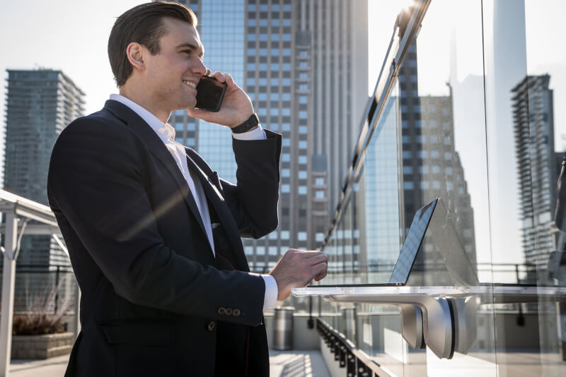 Alumnus Jason Growhowski stands in front of a wall-mounted desk unit while wearing a suit in a big city holding a phone.