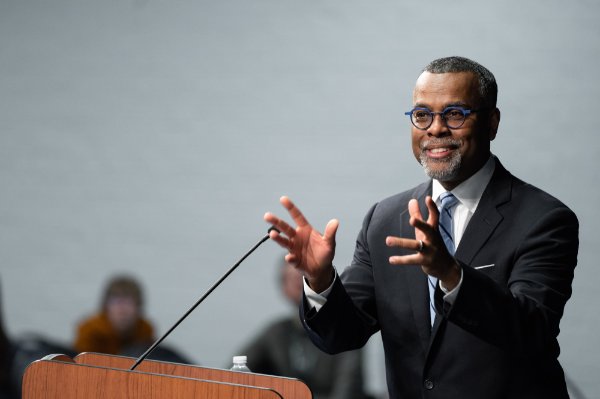Eddie Glaude Jr. at podium with hands raised to audience; he is wearing a dark suit and blue framed glasses