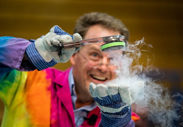  A scientist in a brightly colored lab coat lifts a magnet off his hand with another object as steam flows out of his hand. 
