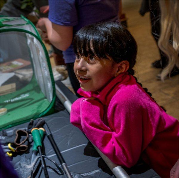 A young girl in a bright pink fleece leans over a table display and looks up with excitement during a hands-on educational activity.