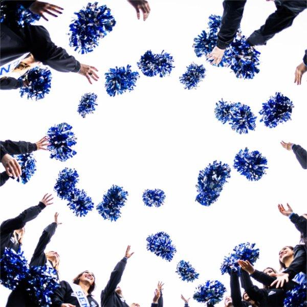 Dancers in black uniforms stand in a circle and toss blue pom-poms into the air against a bright sky.