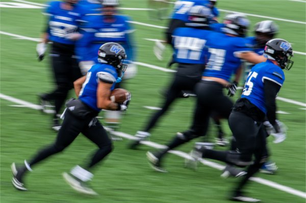  A football player wearing a blue jersey holds a football while running amongst teammates.