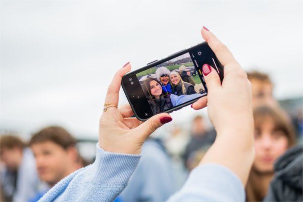  Hands holding  a smartphone show a selfie with three people at an outdoor event.