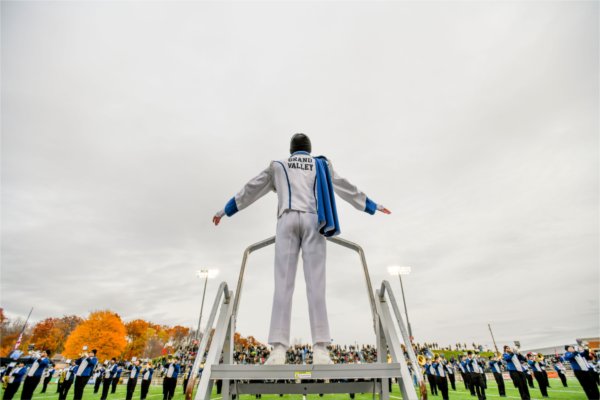  A marching band conductor stands on a platform overseeing band members on a field with autumn trees and a crowd in the background.