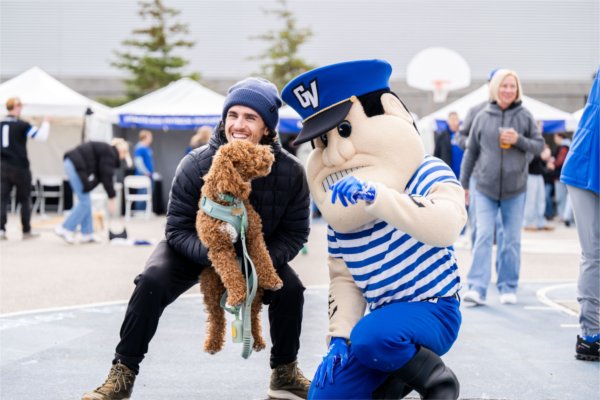  A person holds a fluffy brown dog next to a kneeling mascot in blue and white attire.