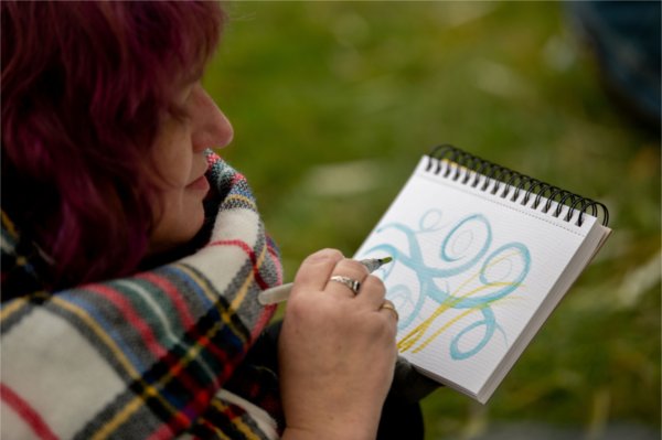  A person wearing a plaid scarf paints abstract patterns in a spiral notebook.