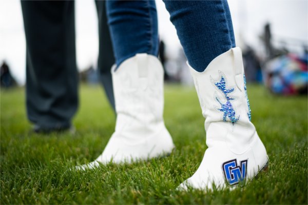  Close-up of white cowboy boots with blue embellishments and a &#8220;GV&#8221; logo, worn over blue jeans, standing on grass.