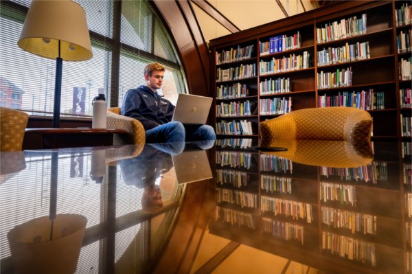  A student works on a laptop in a library with bookshelves and a large window in the background.