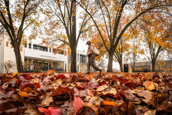 A person wearing headphones walks past the Lake Michigan Hall building with autumn leaves on the ground.