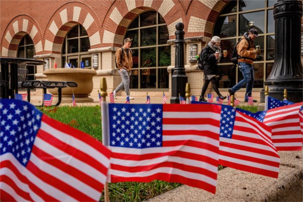 Small American flags are planted in the grass while people walk in front of a brick building in the background.