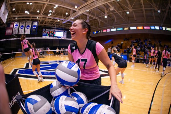 A volleyball player in a pink jersey laughs next to a basket of volleyballs during an indoor match.