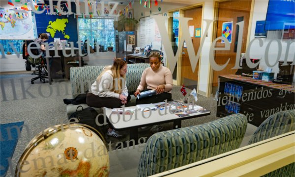  Two people seen through a glass window inscribed with &#8220;Welcome&#8221; in multiple languages sit on a couch inside an office with maps, flags, and brochures.