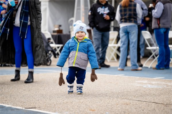  A child in a blue and gray jacket and beanie walks on the pavement while brown mittens dangle from their coat. 