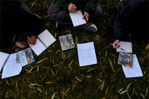 Three people sit on the grass with watercolor palettes and spiral notebooks open.