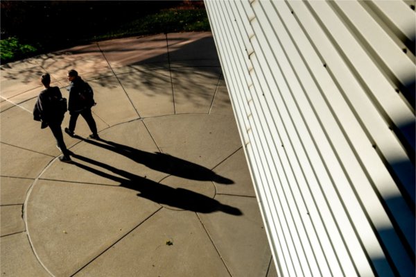  Two people walk on a circular concrete path with long shadows, next to a white slatted structure.