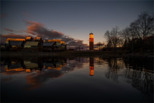  A scenic view of campus at dawn with a building and clock tower reflected on the surface of water.