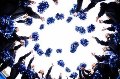Dancers in black uniforms stand in a circle and toss blue pom-poms into the air against a bright sky.