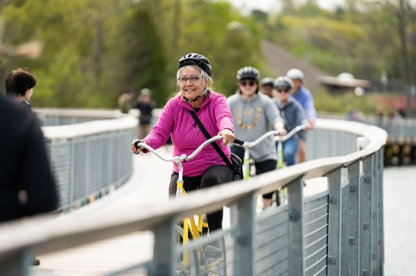 A curve of people on bikes, the front person smiling, ride along a lake on a railed boardwalk. 
