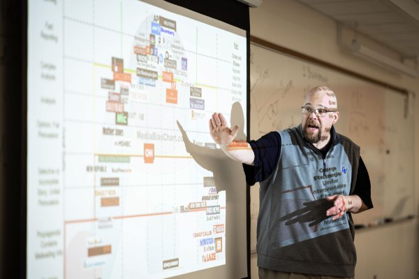 A professor gestures toward a projector screen while teaching a class. Part of the projection is falling over his face and body in addition to the screen. 