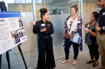 A person gestures toward a poster board while talking to others.