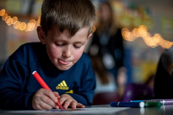  A young boy determinedly colors a photo of a cupcake with a red pen. There are lights strung up in the back of the classroom.