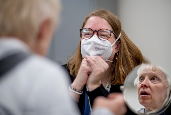  A person wearing a mask sits across from a person who is reflected in a round mirror while testing out her hearing aids. 