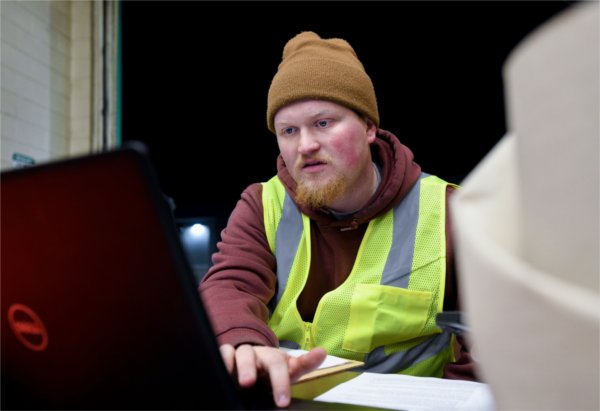 A person wearing a yellow work vest and beige stocking cap works at a computer.