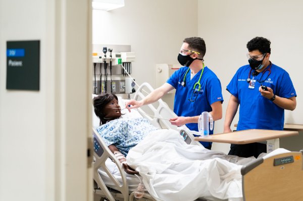 two nursing students stand at the bedside of a sim patient in the lab