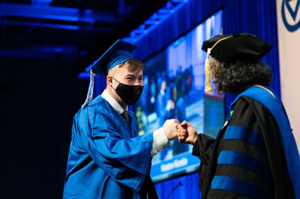 A graduate in a cap and gown fist bumps with a dean.