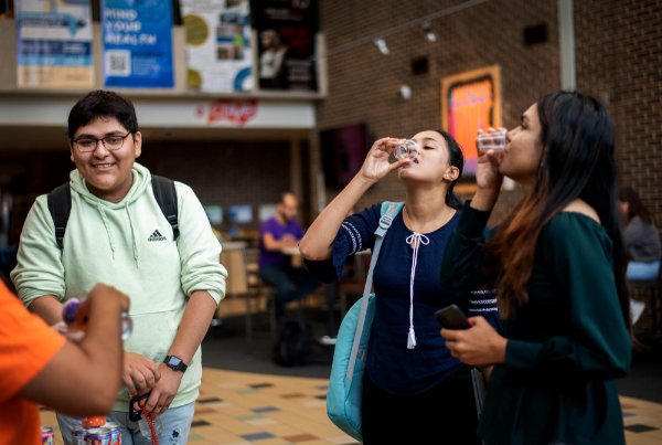 Two students at right drink coffee samples from a plastic cup, student at left in green shirt and backpack smiles at woman handing out samples