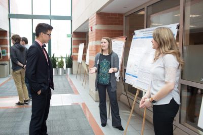 A Student Scholars Day participant stands next to a poster explaining it to another person. A person in a white shirt looks on at right.