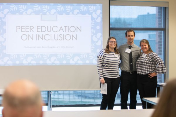 three people standing next to projection screen that reads Peer Education on Inclusion