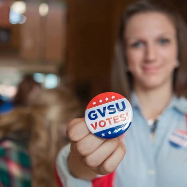 A photo of a student holding up a sticker that says "GVSU Votes!"